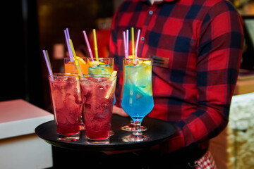 The waiter holds a tray with Multicolored alcoholic and non-alcoholic cocktails with ice and straws