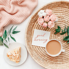 Wicker tray, cup of coffee, piece of cake, rose flowers, eucalyptus branch, pink knitted plaid or blanket, card with text GOOD MORNING. Breakfast in bed. Stylish home interior decor. Flat lay.