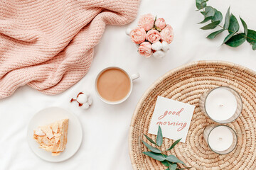 Obraz na płótnie Canvas Wicker tray, cup of coffee, piece of cake, rose flowers, eucalyptus branch, candles, pink knitted plaid or blanket, card with text GOOD MORNING. Breakfast in bed. Stylish home interior decor. Top view