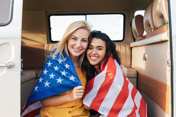 Two young female friends with American flag sitting in a camper van. Beautiful women with USA flag...