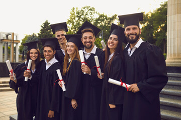 Group of happy graduate students in graduation gowns posing near university campus. Young people...