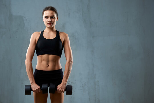 Front view of muscular pretty woman wearing black sports underwear and carrying dumbbells. Isolated portrait of pumped fitnesswoman posing on gray studio background, loft interior. Concept of sport.