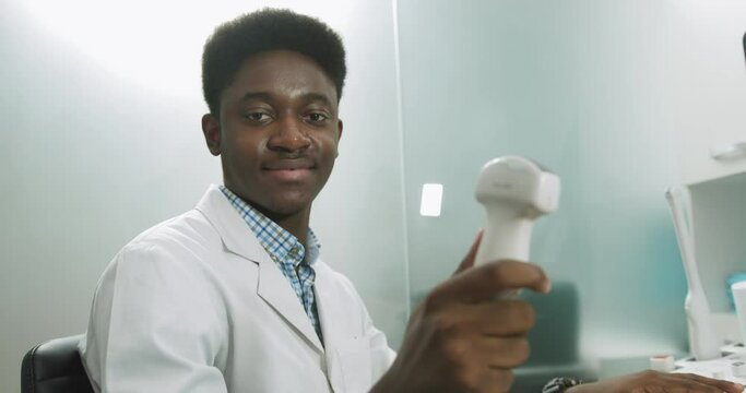 Smiling African man doctor with ultrasound transducer in hand, working on modern ultrasound scanning machine in light room in clinic. Portrait Of 4D Ultrasound Scanning Machine Operator