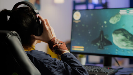 Focused woman gamer sitting on desk, putting on headset with a mic and starts playing space shooter video game. Online streaming cyber performing on powerful personal computer during gaming tournament