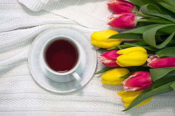 Cup of tea in porcelain cup with bouquet of flowers yellow and pink tulips on white fabric. Gift of surprise breakfast on special occasion. Positive start of a day, good morning concept.