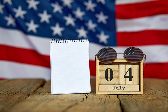 Wooden calendar with numbers on which the image of the date of American Independence Day on a wooden background and glasses with a flag, as well as a blank notebook. Independence