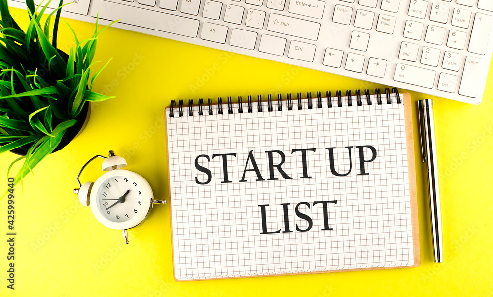 Poster start up list text on notebook with keyboard , pen and alarm clock on the yellow background