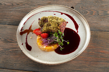 Top view of beef meat with pistachio topping served with cranberry sauce. Tasty meal with fruits, berries and fresh sprouts presented on ceramic white plate in luxurious restaurant, wooden table.