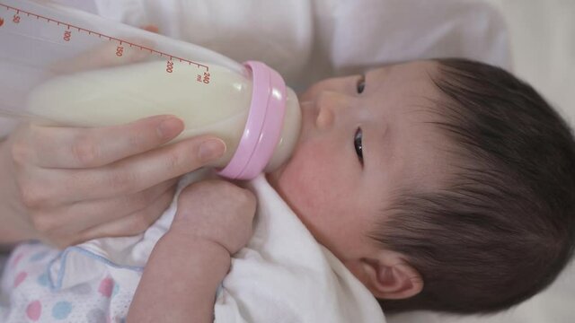 asian innocent new baby held by her mother is getting sleepy while suckling milk from the bottle in the bedroom.
