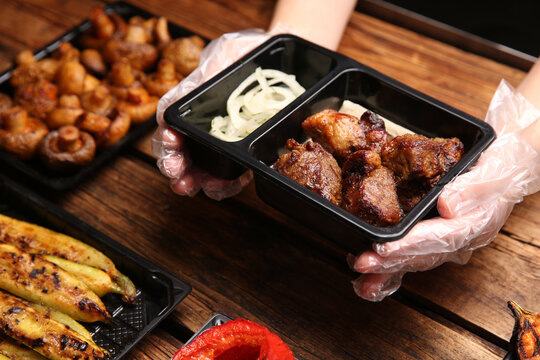 Waiter With Plastic Container Of Tasty Shish Kebab At Wooden Table, Closeup. Food Delivery Service