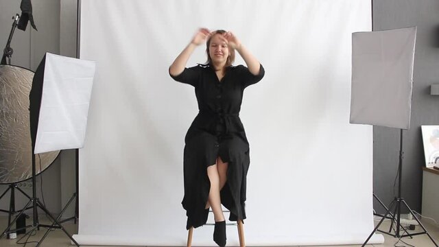 photo studio with white background. girl in black dress is sitting on the high chair and smiling cute. funny conversation with model before shooting. making off interview. lifestyle concept