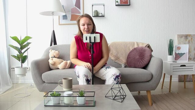 young girl in white pants and pink shirt is sitting casual on the gray sofa and talking on the mobile phone on the tripod for phone in light room at home in the morning. lifestyle concept, free space