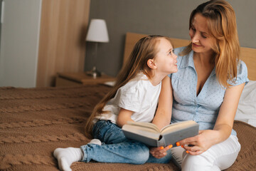 Portrait of young mother and adorable daughter reading children book together sitting on bed in...