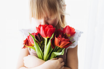 Smiling small girl holding and sniffing bouquet of red tulips. Greeting card