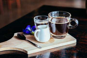 A cup of black coffee with a white small jar and glass of water on wooden small board