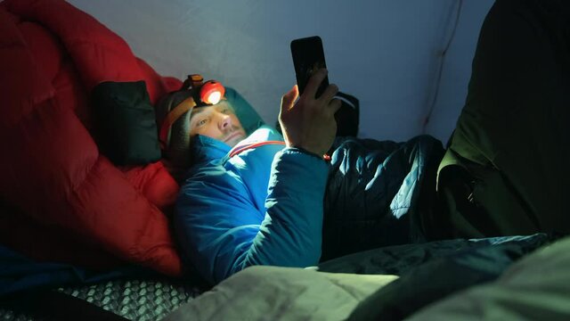 A tourist lies in a tent in winter with a flashlight and warm clothes and looks with a swipe at the phone.
A man lies in a cold tent in winter, he has steam from warm breath.