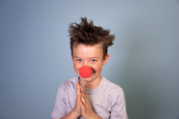 cool young schoolboy with wild hair posing with red clown nose on blue background and is happy