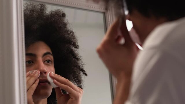 Male facial care. Skin cleansing. Hygiene routine. Confident handsome guy with long curly hair applying toner with cotton pad looking in mirror.