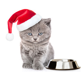 Cute kitten wearing red santa hat sits with empty bowl. isolated on white background