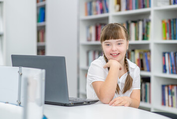 Fototapeta na wymiar Young girl with syndrome down uses a laptop at library. Education for disabled children concept