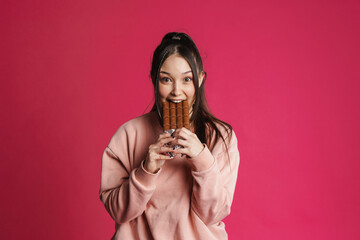 Happy young woman eating chocolate bar over pink