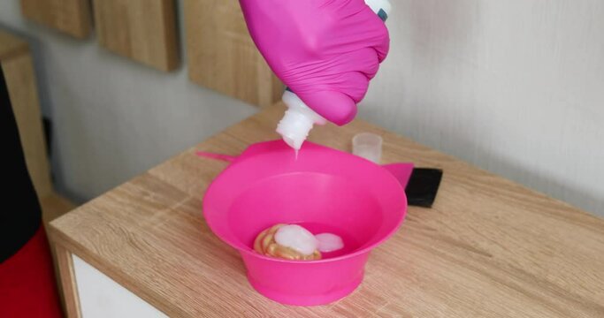 Unrecognizable female wpman in pink glove squeezing pigment in bowl while preparing to dye hair at home.