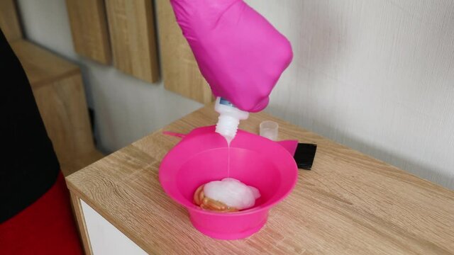 Slow motion Unrecognizable female wpman in pink glove squeezing pigment in bowl while preparing to dye hair at home.