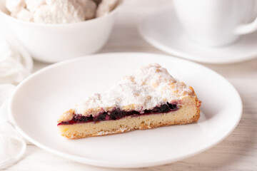 A piece of handmade grated cherry pie on a white plate. Sprinkled on top with powdered sugar. White wood background. Close-up. In the background a white cup and white cookies in a bowl 