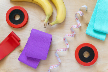 Fitness flat lay. Dumbbells, rubber bands for sports, bananas and centimeter on wooden background