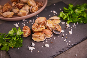 Fresh mussels in a vangole shell are piled on a black slate tray on a dark gray table, surrounded by parsley leaves and sea salt. The concept of healthy food and fresh seafood and delicacies.
