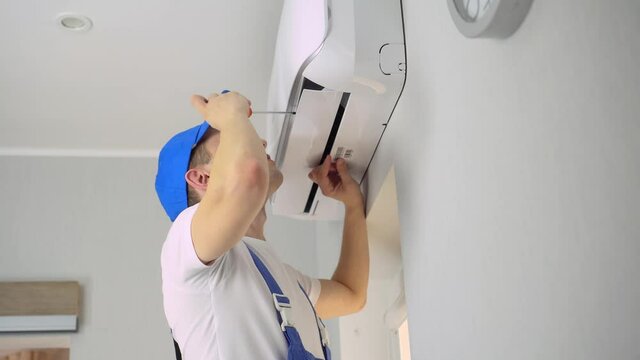 A young foreman in uniform, blue overalls and cap, white T-shirt, installs, repairs and does maintenance of the air conditioner in the apartment room.