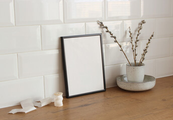 Spring still life. Blank picture frame mockup on wooden table background. Easter composition with blooming goat willow, pussy willow or great sallow in a vase and white silk ribbon. 