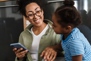 Black smiling woman playing her daughter while using mobile phone
