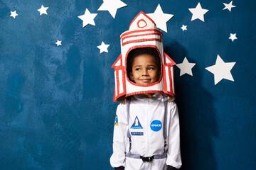African-american boy playing astronaut with handcrafted rocket helmet on blue studio backdrop with white stars. Portrait of smiling mixed race child dreaming of cosmic space. Childhood and imagination