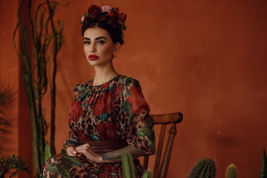 Portrait of beautiful dark-haired tattooed woman with crown braid hairstyle and floral head wreath wearing chic chiffon dress sitting on the wooden chair among cactuses. Mexican style. 