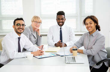 Multy-ethnic group of business people working while sitting at the office desk and smiling at camera