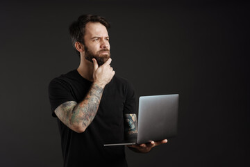 Bearded brunette man with tattoo frowning and using laptop