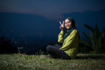 Pretty girl sitting on grass, relaxing, enjoying Kuala Lumpur city view during sunset. Portrait of young Asian woman, wearing hijab and green attire.