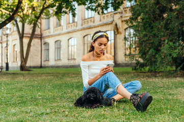 Beautiful woman sitting on the grass in the park with a little black dog and enjoys the Internet on a smartphone with a serious face.