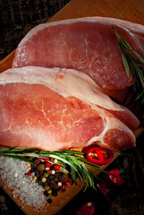 Raw steak on a wooden board on a black background. Red meat on an old shabby table. Contrasting dramatic light as an artistic effect. Fresh pepper, salt and rosemary near the meat.