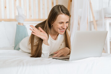 Obraz na płótnie Canvas Freelance woman having a video call chat, typing at laptop and online shopping, lying on the white bed. Happy relaxed girl woking from home office. Distance learning online education and work.