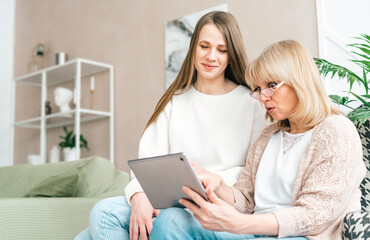 Mature surprised senior woman with smiling adult daughter using digital tablet computer device. Caucasian mother and daughter sitting on armchair at home interior in background. Technology concept.