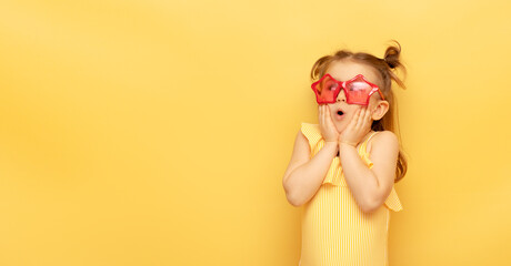 Little child girl in striped swimsuit and red funny summer sunglasses surprised expression looks at camera posing on yellow background, studio portrait.Advertising of children's products and sale