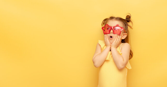 Naklejki Little child girl in striped swimsuit and red funny summer sunglasses surprised expression looks at camera posing on yellow background, studio portrait.Advertising of children's products and sale