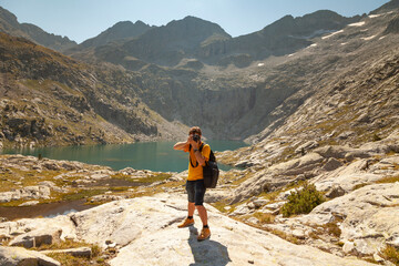A hiker takes photos of the natural landscape with a reflex camera, in a lake with turquoise waters that reflect the mountains in summer, Aragonese Pyrenees, Huesca, Posets-Maladeta Natural Park.