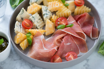 Round grey plate with jamon, various cheese, cherry tomatoes and green basil leaves, selective focus, close-up