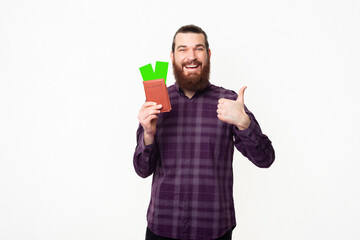 Joyful handsome young bearded man wearing checkered shirt showing thumb up and passport with tickets