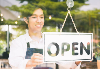 Young asian man Flip the open label to welcome customer and open the coffee shop in morning. Male asian Barista open the coffee cafe at doorway with reflection in mirror.