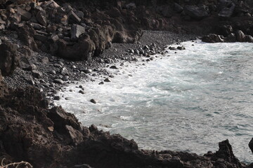 Dark black rock cliff and stones coast with turquoise foamy ocean waves. Side view of ocean water and rocks formed from solidified lava of volcano and pebbles.