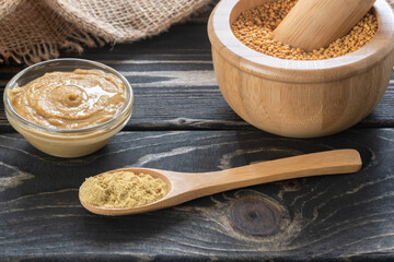 Mustard seeds in a mortar, ground mustard seeds in the spoon and mustard dip in the bowl on brown wooden table